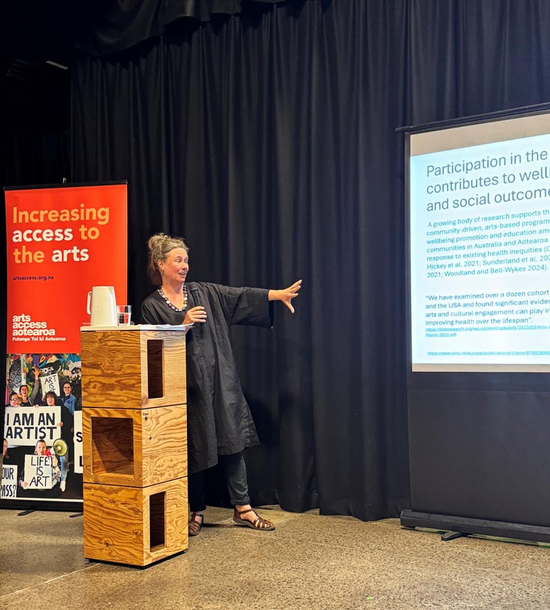 Dr Molly Mullen stands behind a lectern, pointing with her left arm at her powerpoint presentation 